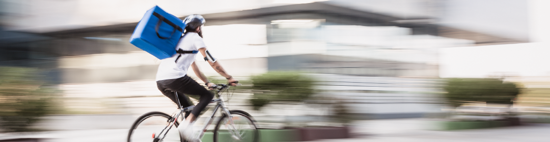 a man quickly riding a bicycle down a sunny street while wearing a blue delivery box on his back